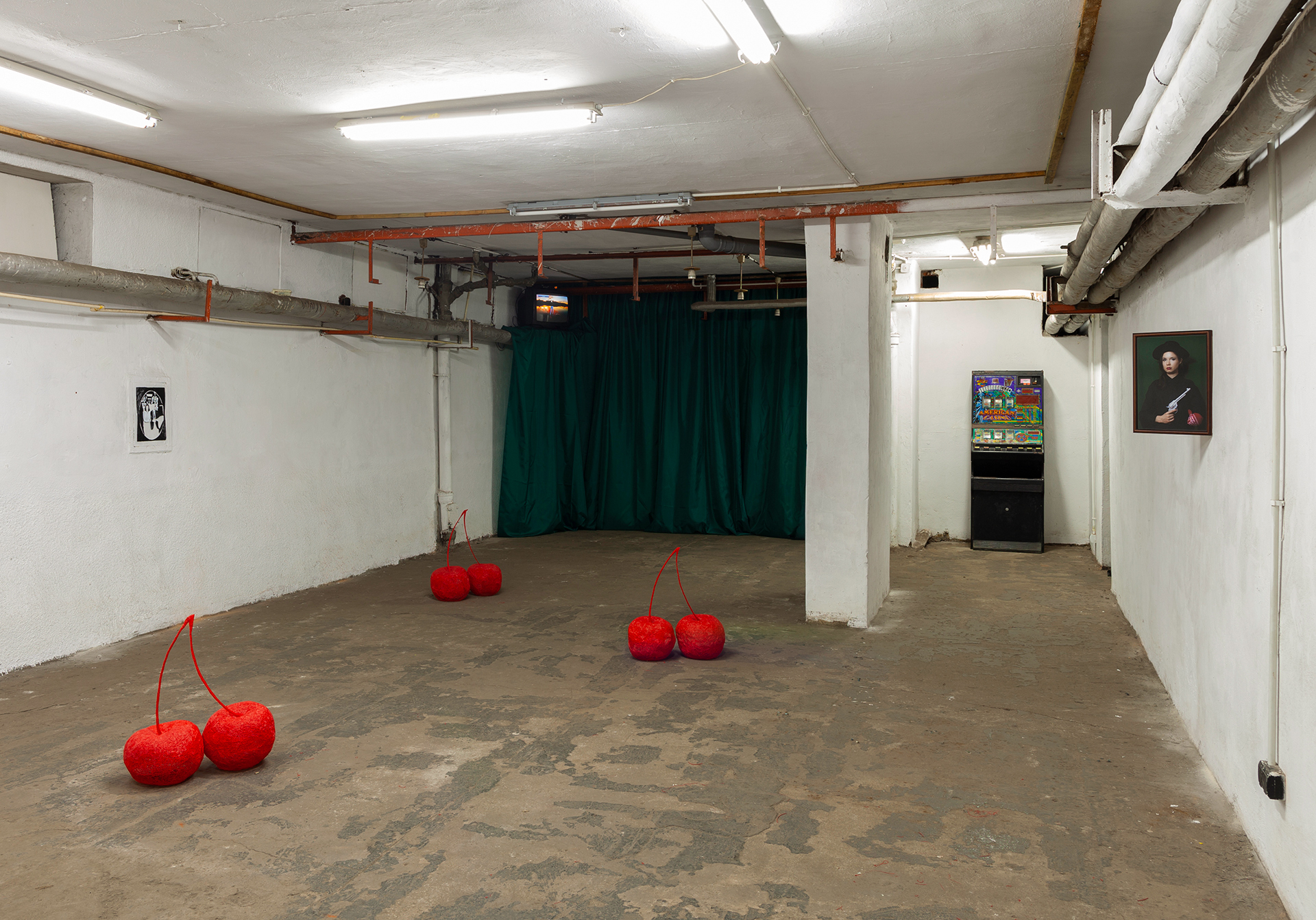 In the image there is an interior in a basement with no windows. Scattered through the middle of the space are three sets of oversized red cherries, much like the ones in slot machines. Behind them, towards the back of the room is a dark green curtain. At the top left corner of the curtain is a TV, presenting a video containing a sunset. Yet the TV is far away from the camera and small. On the right side of the curtain, against a white wall is a slot machine. On the wall next to the machine, the one that in perspective is coming closer to the camera, somewhere mid-way across the space is a photo of a white woman dressed in a black hat, holding a revolver, with a red bandana on the end of her arm - around the elbow. She is portrayed on a dark green background. Across the room, on the wall on the other side of the cherries, there is a small black and white image of a couple. The room has additional visible elements typical for basement spaces such as pipes crossing the room near the ceiling.
