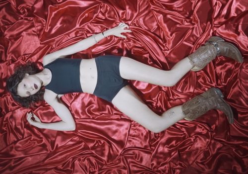 Overhead shot of a trans* non-binary cowboy wearing a black binder, underwear and brown cowboy boots, lying on red satin and looking into the camera