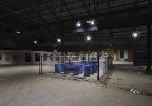Four rows of blue plastic barrels are surrounded by a chain link fence with barbed wire. They are in a large warehouse-like space with concrete floors.