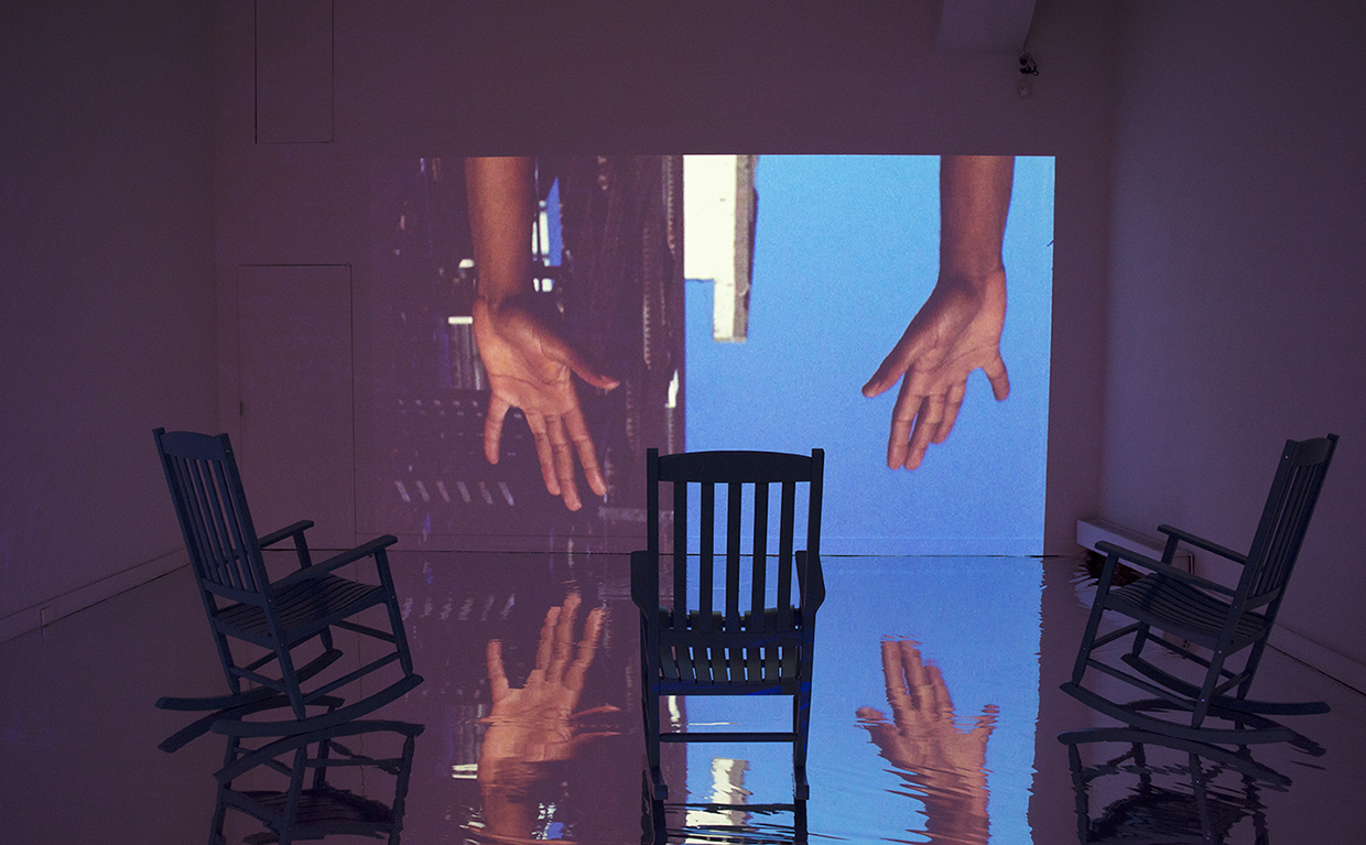A video projection of brown hands reaching downwards in front of a blue sky and sliver of a brick building is reflected on a silver mylar floor with three rocking chairs.