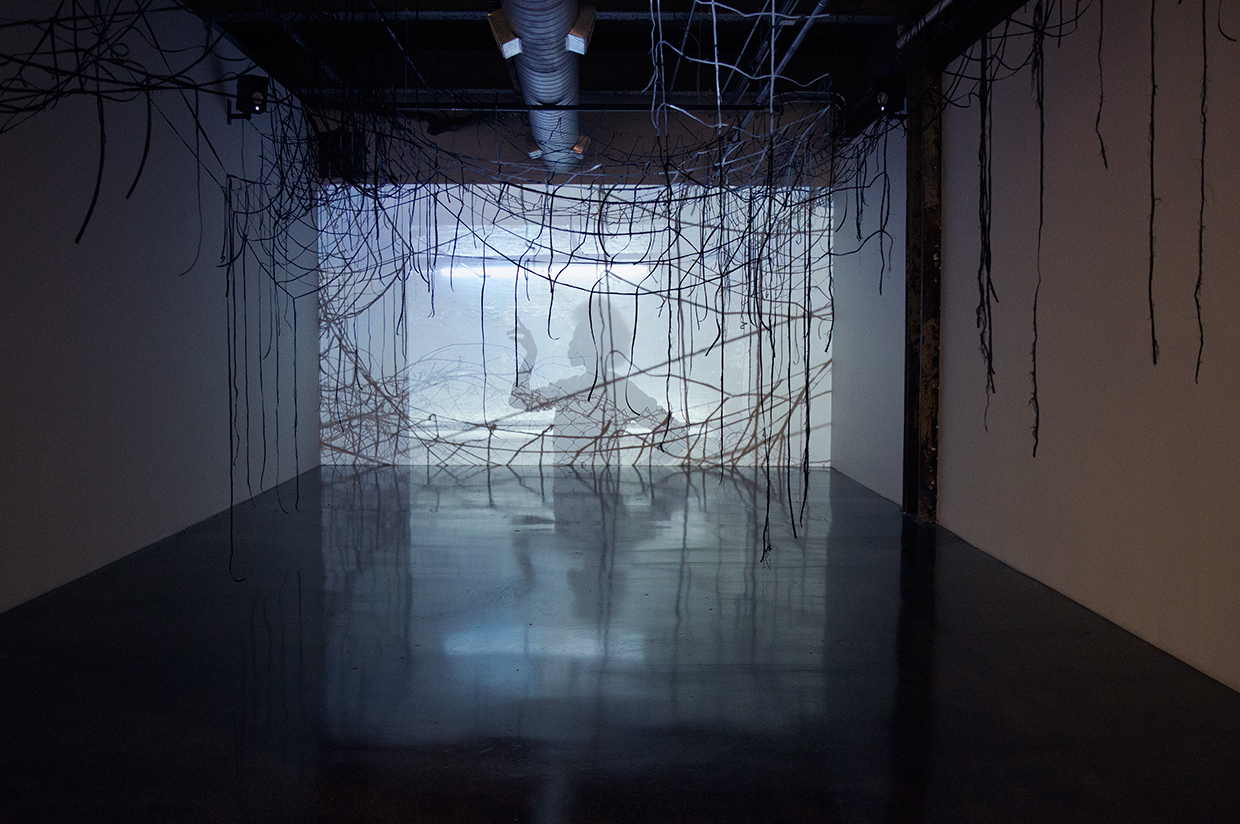 Thin lines of sculptural elastic criss-cross the gallery from wall to ceiling; a video projection through them shows a silhouetted figure with one arm gesturing outwards in hues of blue and gray.