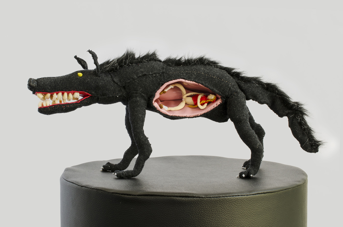A black wolf sewn out of fabric stands on a cylindrical black base. It has human teeth in its mouth and a small woman wearing a red t-shirt in its belly.