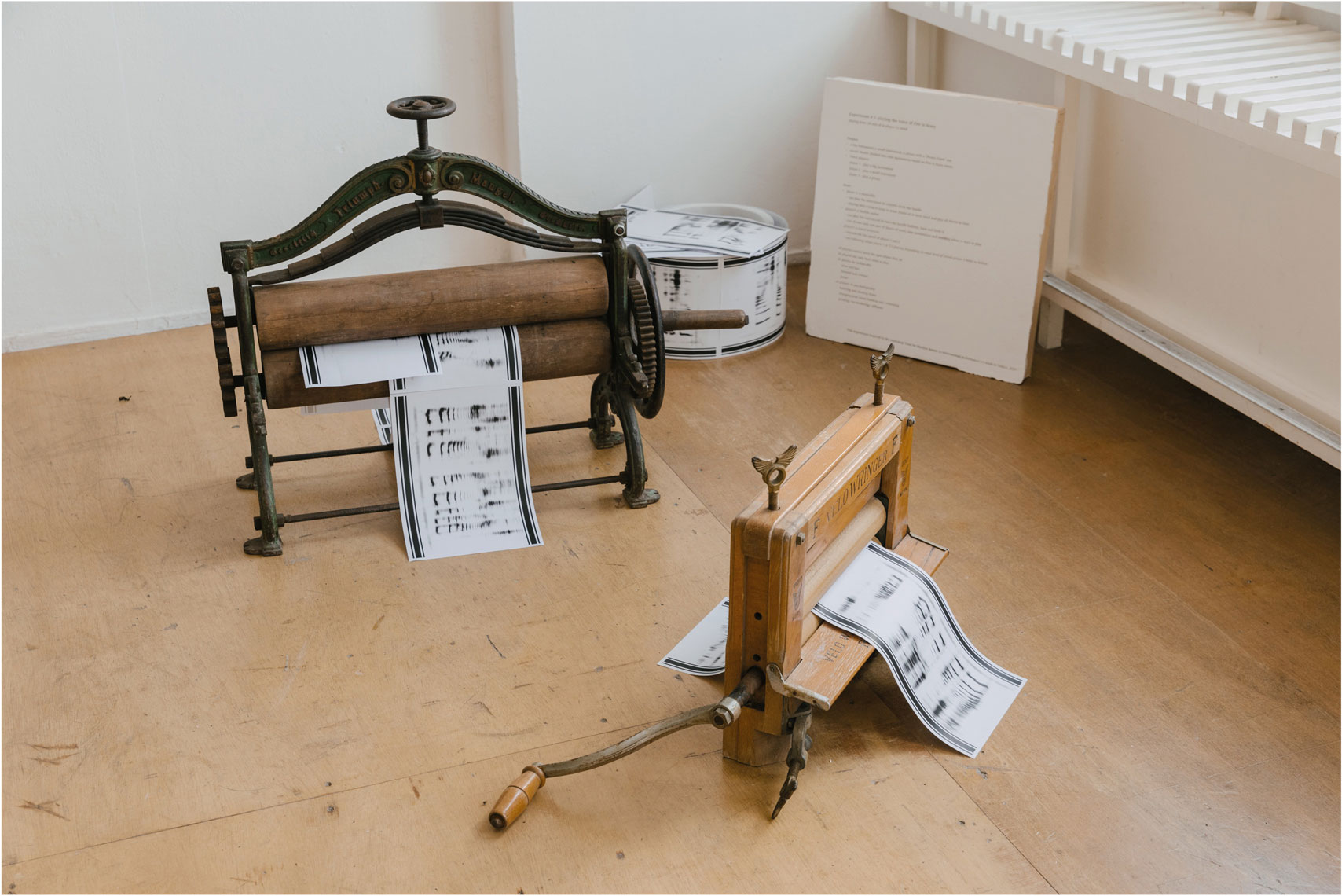 Two old printer parts have two roller bars and one handle to rotate the rollers to make the papers out. Printed spectrograms are installed between the rollers, and leftover spectrograms are connected and shaped paper-roll aside. There is a white wooden panel describing the score for using devices and spectrogram with a phone app.