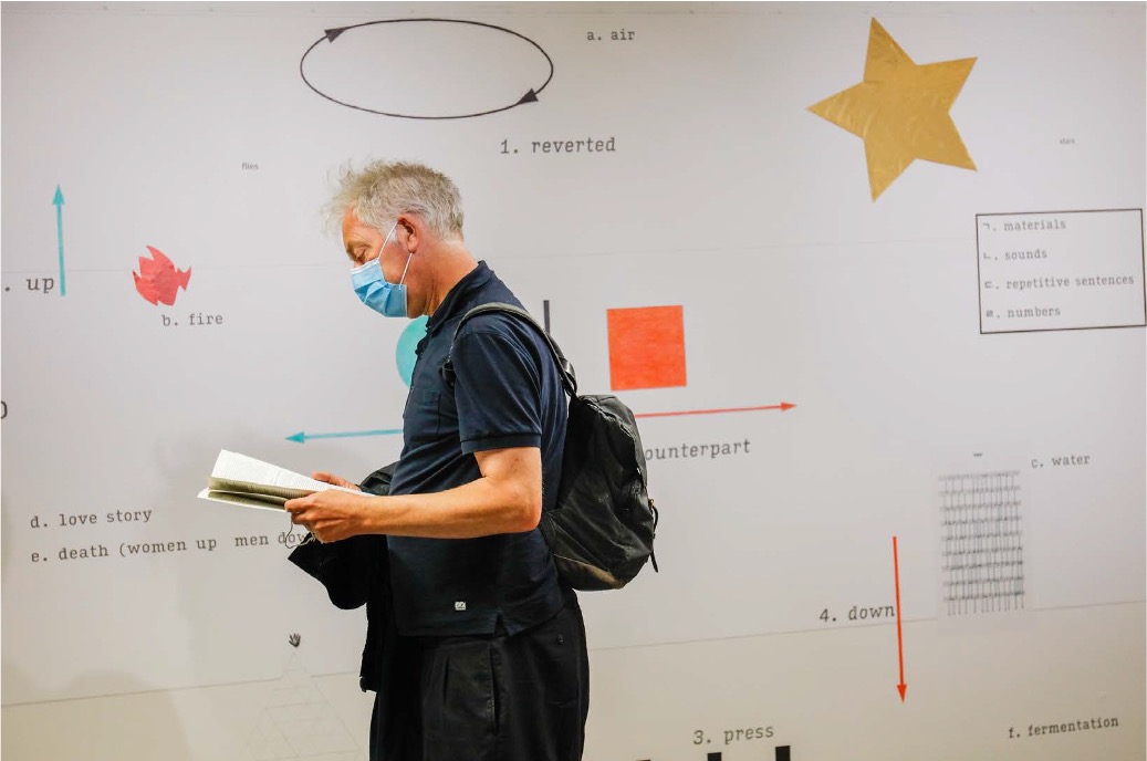The diagram on the wall contains simple shapes, arrows, and text. One visitor is passing by, reading the poster booklet of the project