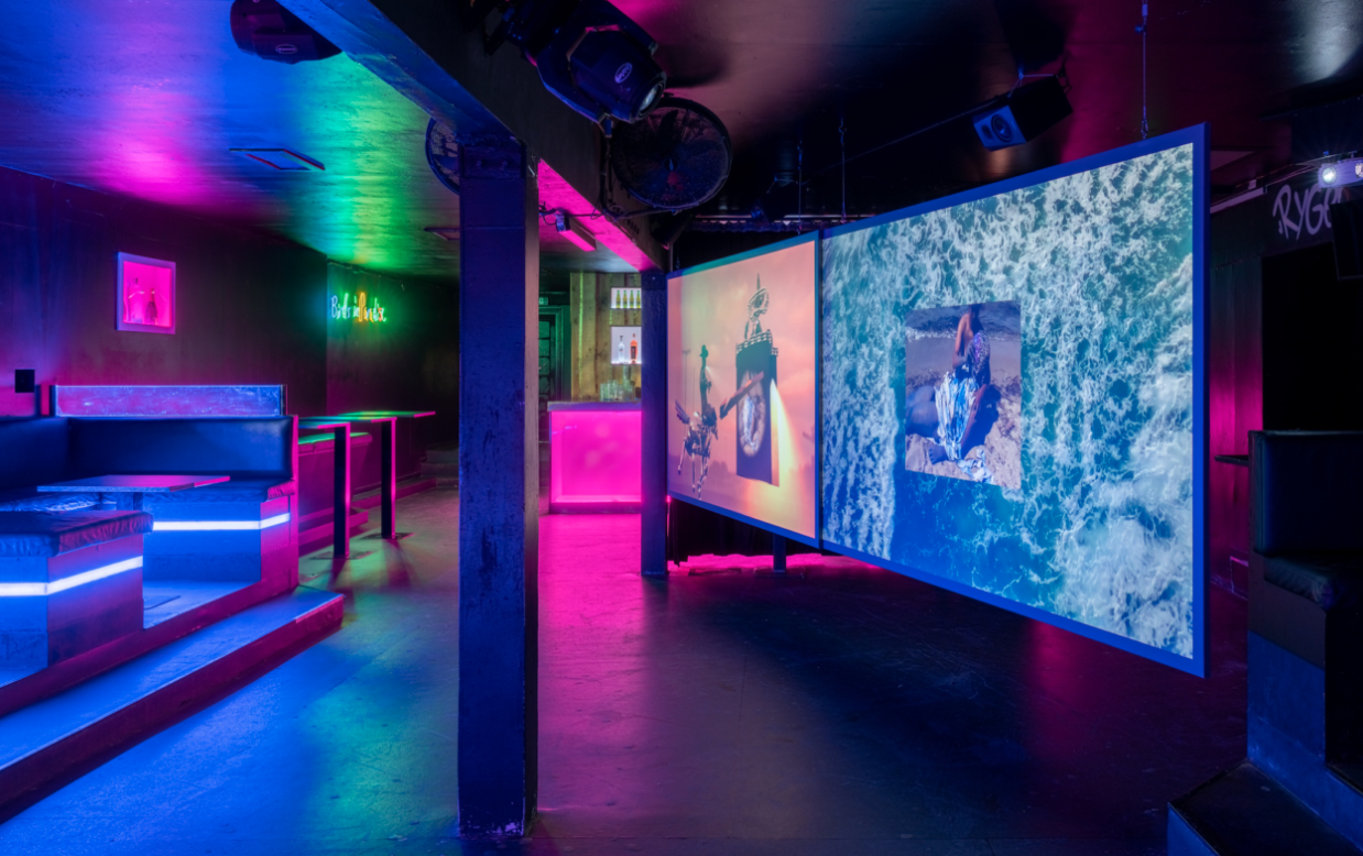 Video projection screens with work by American artist Jacolby Satterwhite installed in a nightclub in Roskilde, Denmark. The lighting is dimmed, pink and blue.