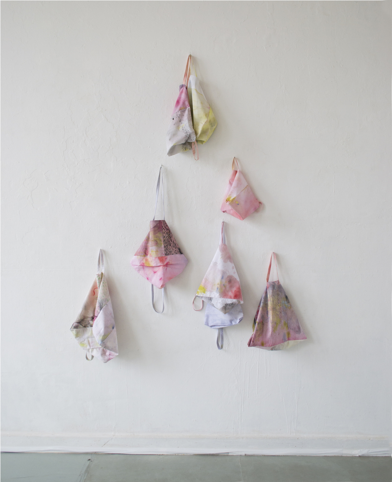 Seven fabric tote bag like forms are installed in a triangle shape on a white wall. The fabric is painted a range of pinks and pastel colours.