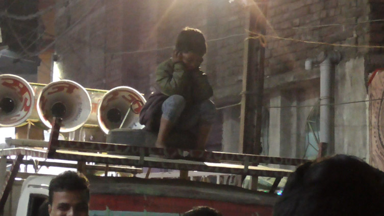A child riding on top of a sound-system-cart (like how one rides an elephant or a camel), covers their ears to protect themselves from the impact of loud-speakers around themselves.