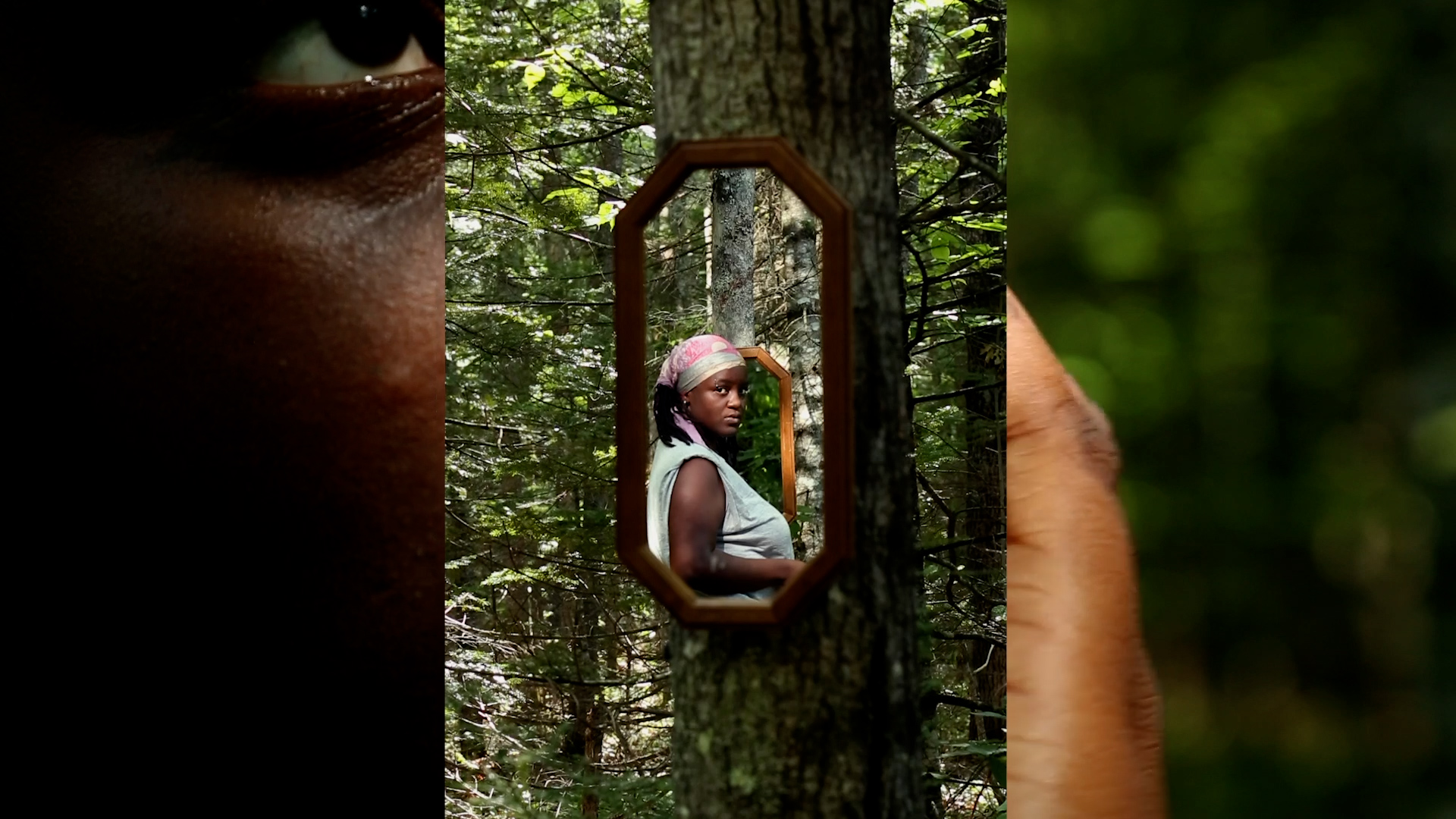 A brown skinned person in a silk bandana turns back to look directly at the camera in a forested area.