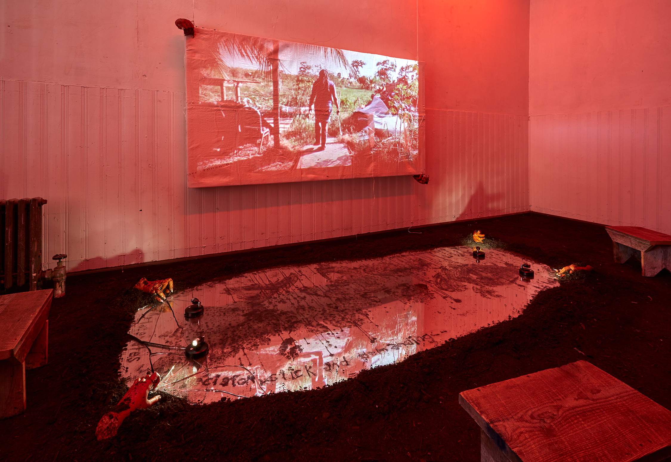 A video is projected in a darkened space awash with a soft red light. The floor is covered in dirt and a cracked mirror.