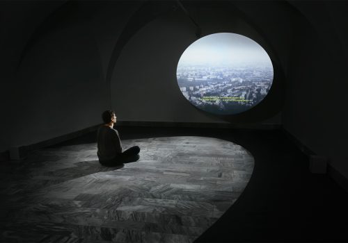 A man sitting in the gallery in front of an ellipse-shaped projection on a marble floor surrounded by a black ellipse-shaped carpet.