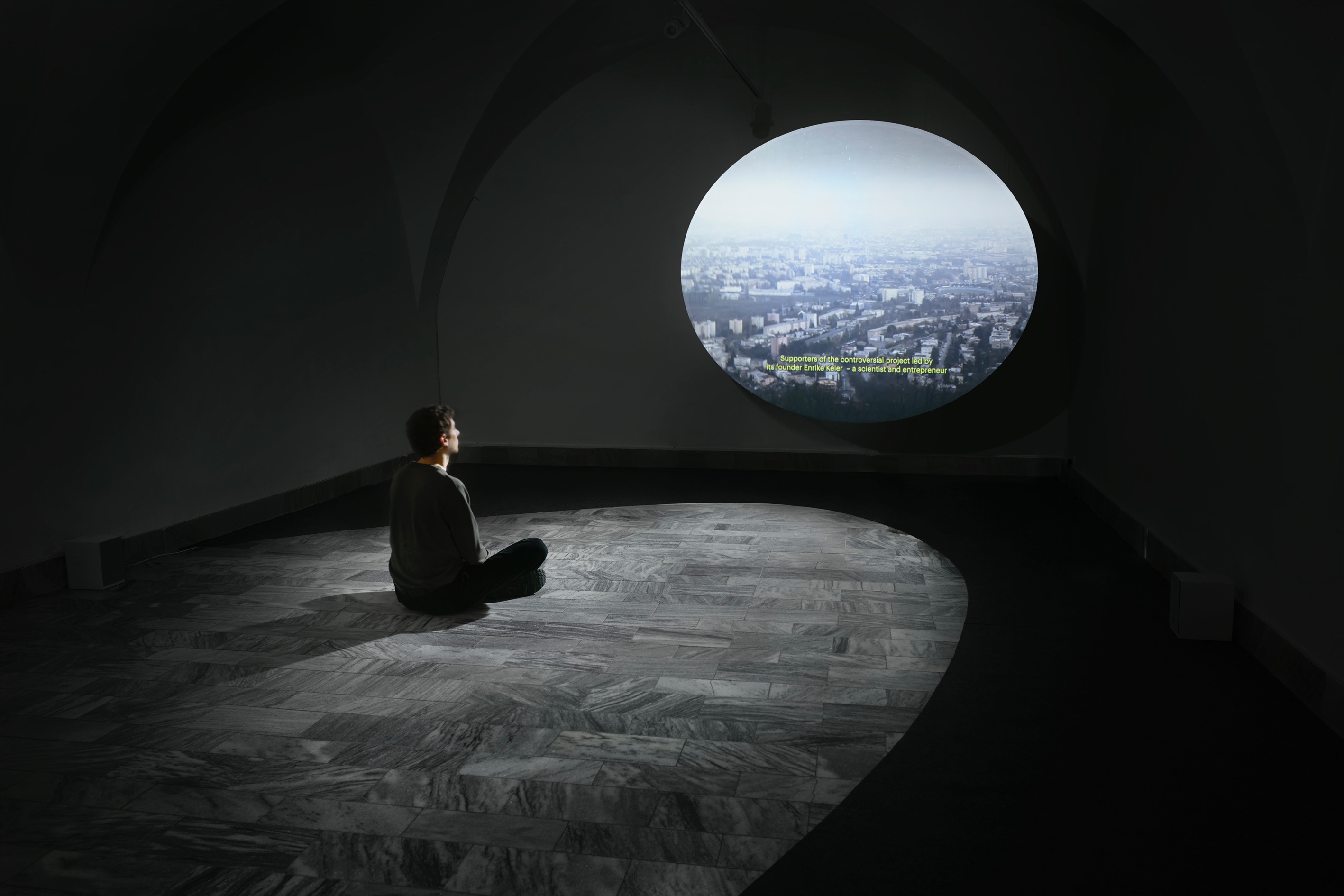 A man sitting in the gallery in front of an ellipse-shaped projection on a marble floor surrounded by a black ellipse-shaped carpet.