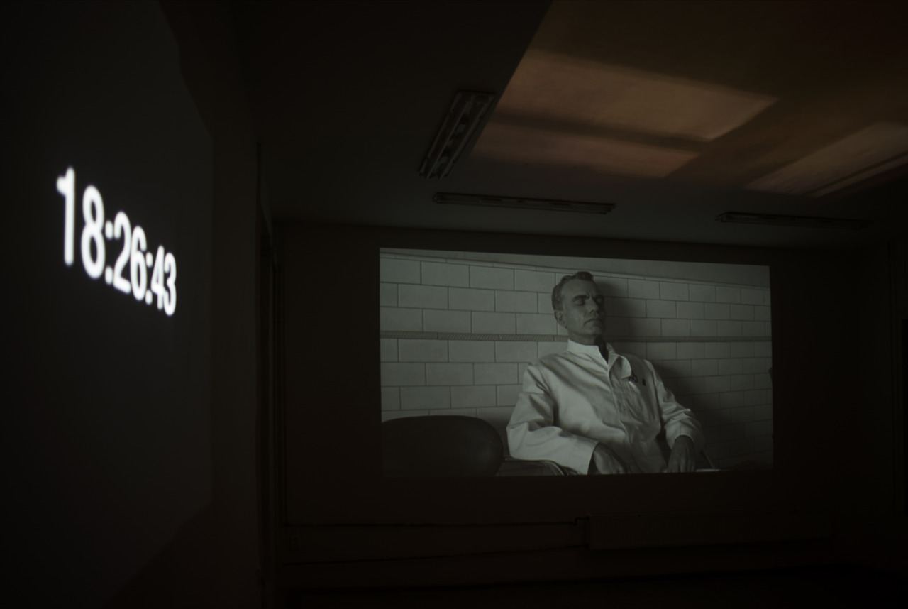 A three-channel video projection in the gallery, on the left wall is a white numerical description of the time in hours, minutes and seconds, on the middle wall is a shot from the film The Man Who Wasn't There where Billy Bob Thornton sits in a white barber's coat and sleeps, on the ceiling is a projection simulating the shadows formed by car lights on the ceiling of the night rooms around the road.