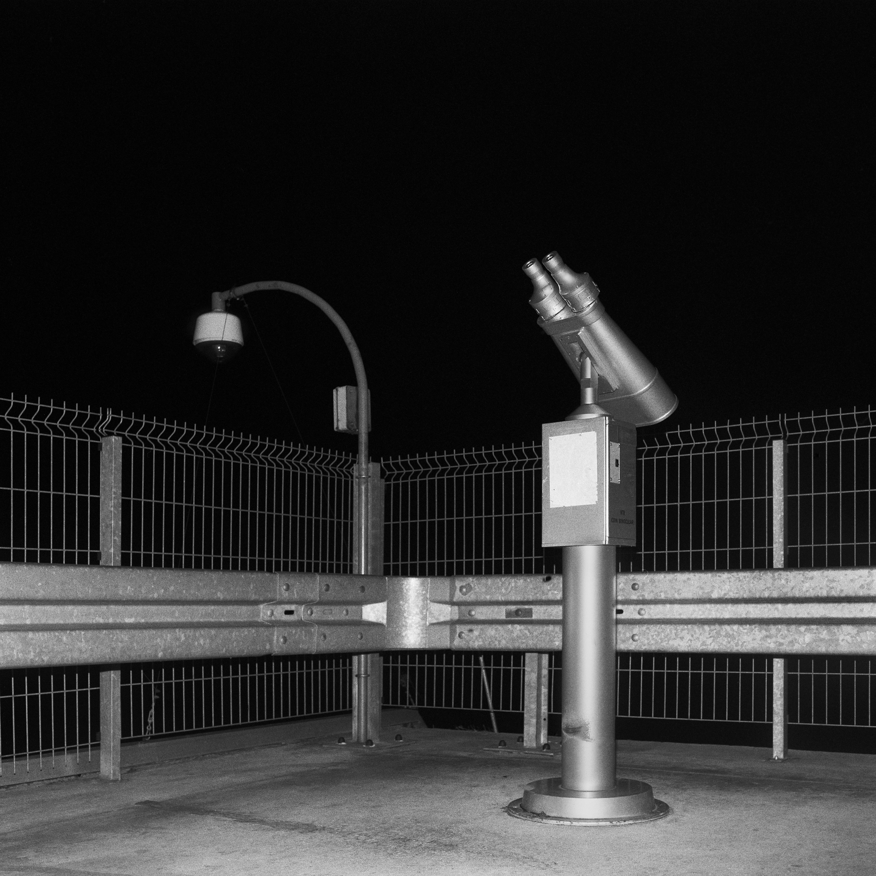 Square black and white photograph at night with flash showing corner of road with bollards, low fence, security camera and metal telescope on base.