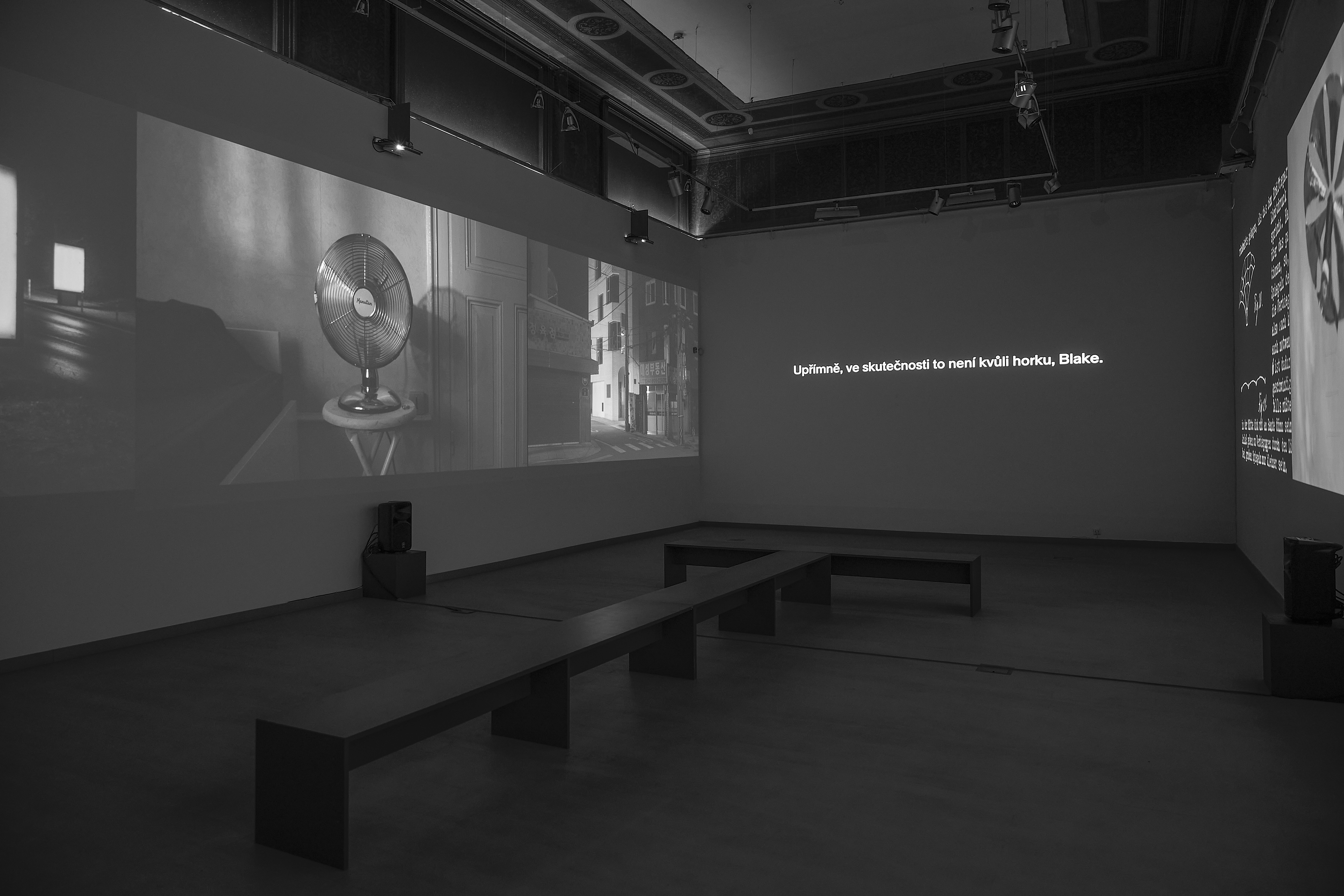 A black and white multi-channel video installation on the gallery walls consisting of a projection of photographs of empty citylights on the left wall, a spinning fan in a night room, a shot of a night street in Seoul, a caption to an audio track on the middle wall, a white graph on a black background on the right, and a shot of a circular satellite with a reflective surface of mirrors.