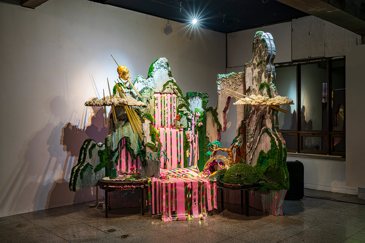 A large, wooden sculpture resembling a landscape sits in a room next to a wall.A pink ribbon is tied around the sculpture. There is a cascading waterfall made of rocks. A green ribbon is wrapped around the base of the waterfall.