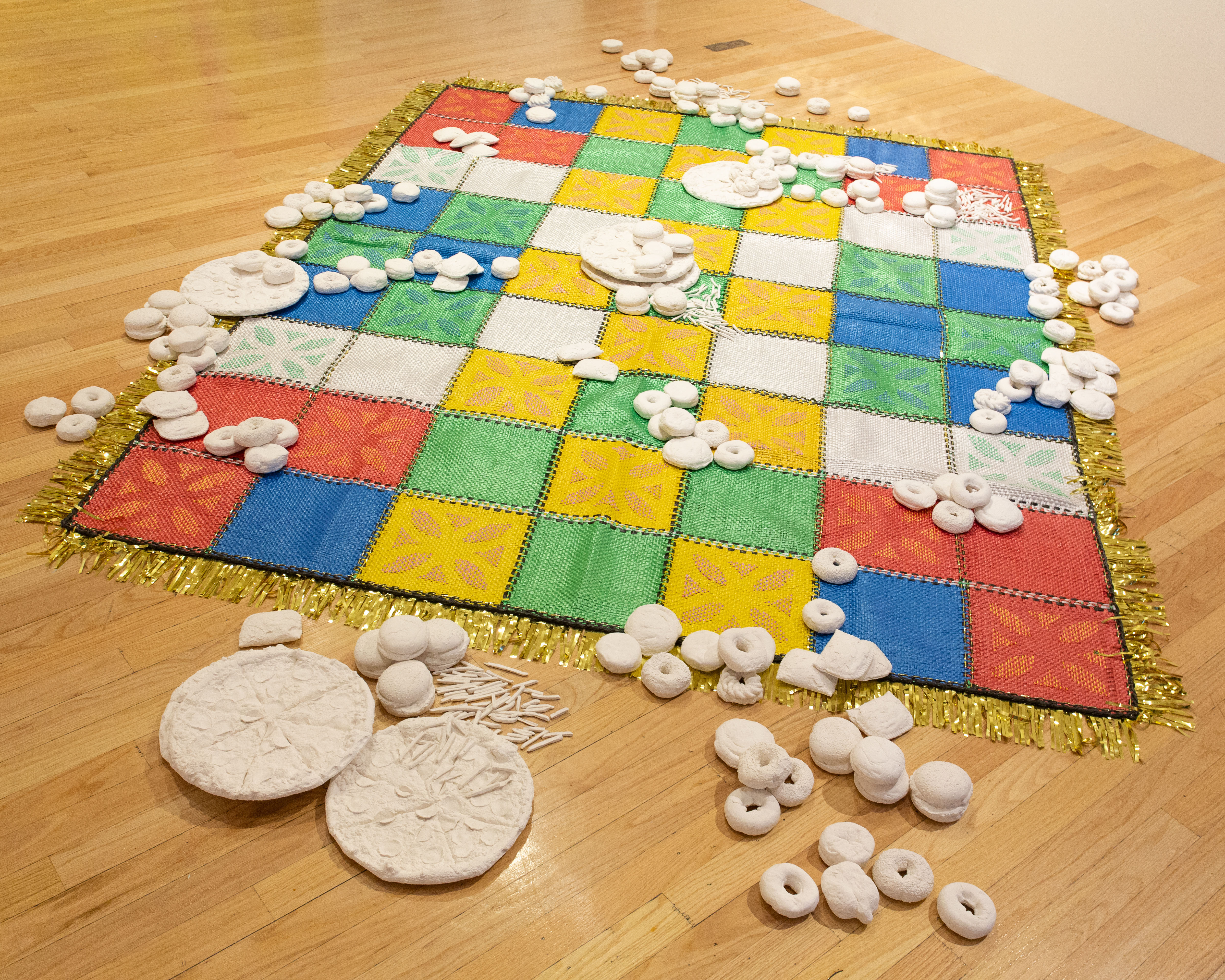 A multi-colored woven mat laid on a wood paneled floor with white plaster casts of fast-food items.