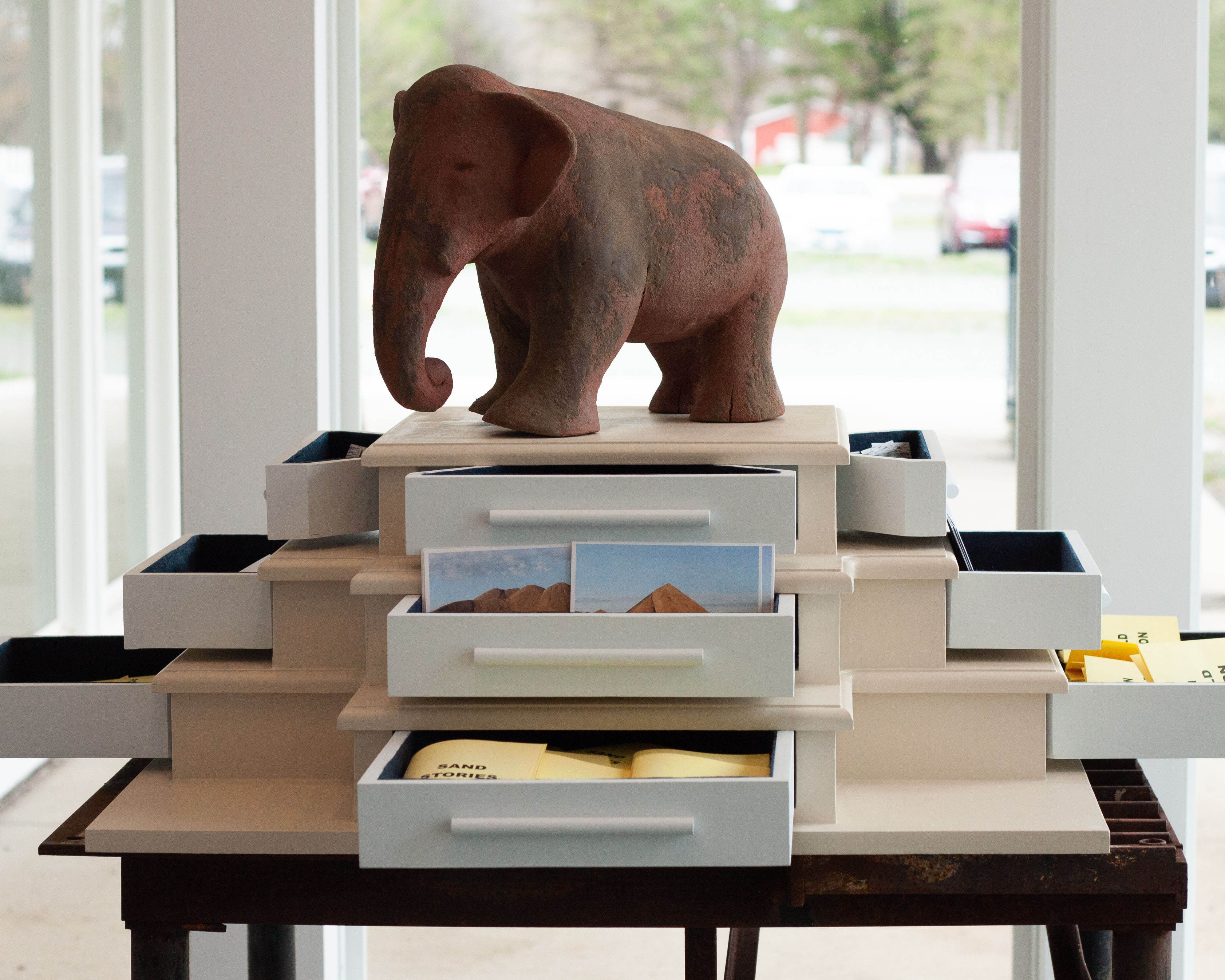 A sculpture of an elephant made with a brown clay covered with patches of sand sits on base with three levels of drawers. The drawers are opened and peeking out are postcards with sandy landscape photos on them, and yellow booklets that are laid flat.