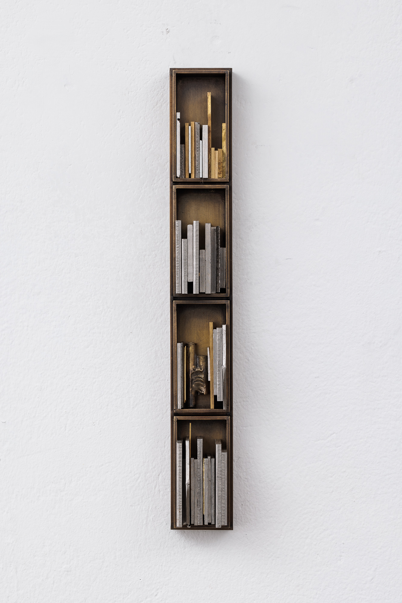 A wall-hanging sculpture made up of four brass boxes in a vertical line. Smaller slivers of metal are squeezed within the boxes.