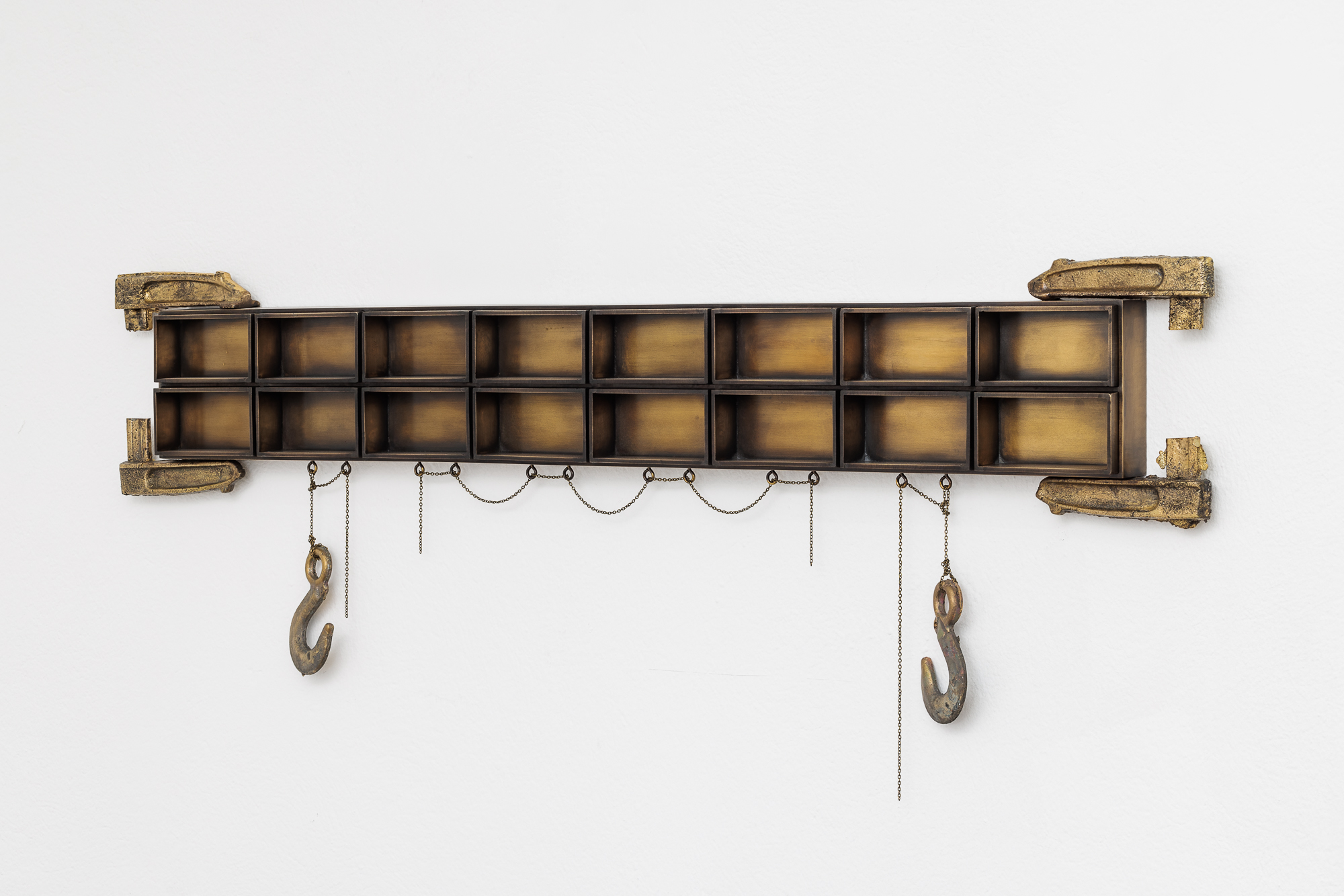 A wall-hanging sculpture made up of two long rows of brass boxes. Cast brass clamps grip the sculpture on the top and bottom. Cast brass hooks hang from the bottom.