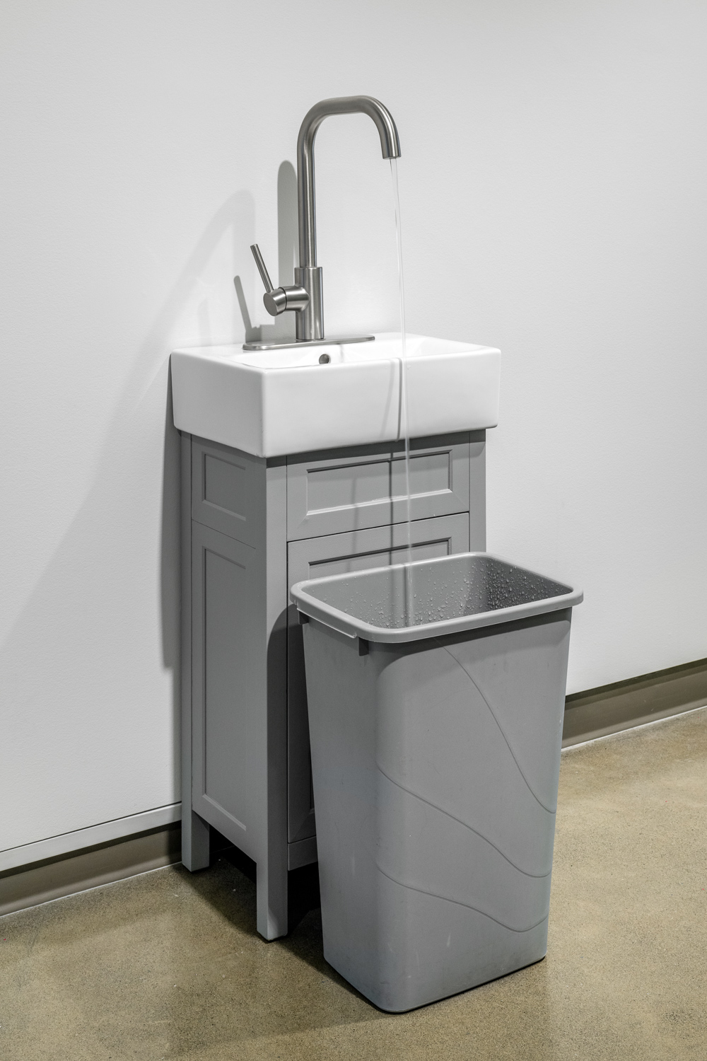 Photo of a water fountain that consists of a sink with an overly large faucet, water pours out of the faucet into a garbage can in front of the sink