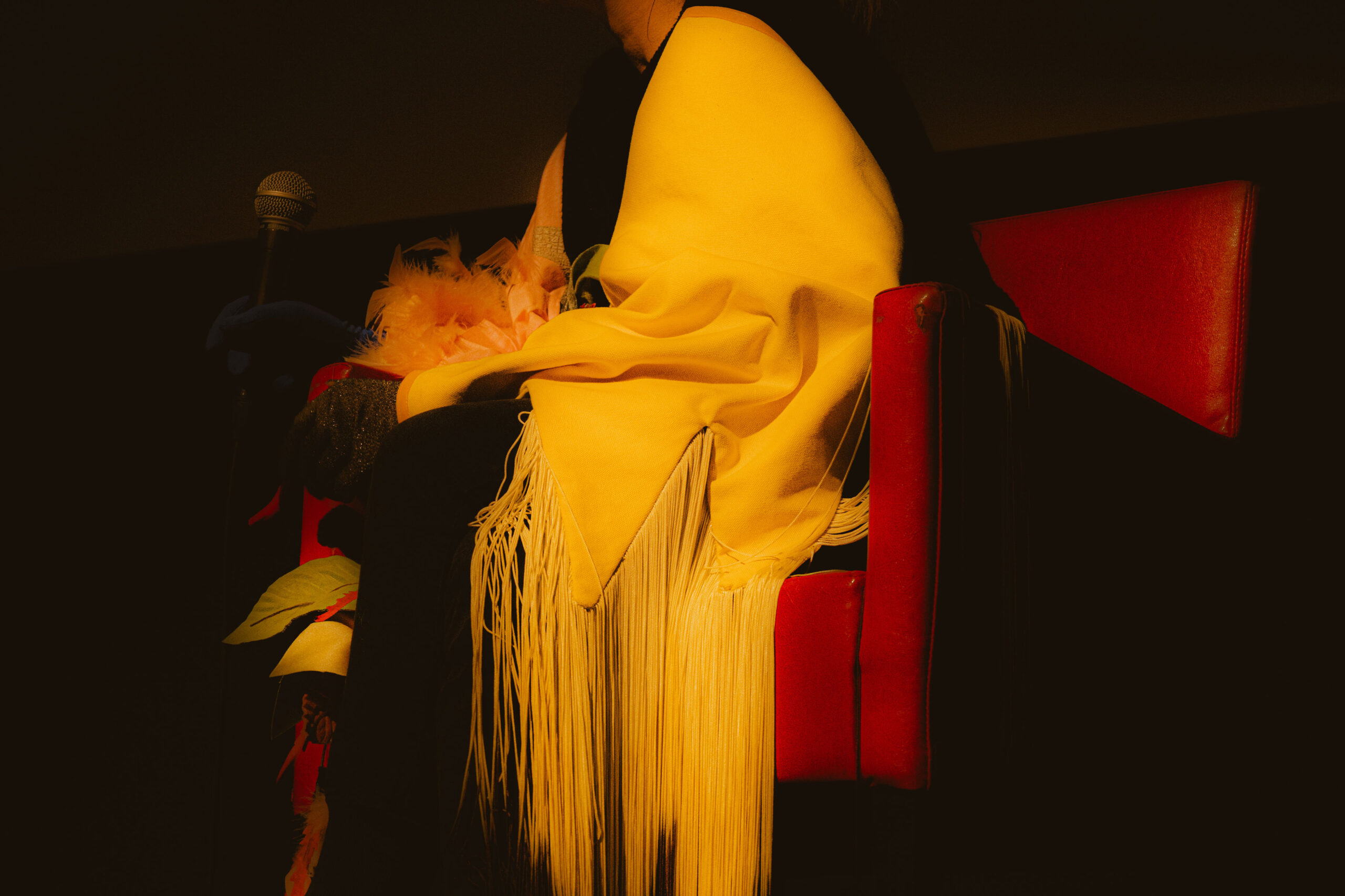 A figure is seated under a spotlight, in a large red velvet chair within a blackened room. The figure’s legs, in black, are crossed with one bent knee perched on the seat. The figure is wrapped in a deep saffron yellow silk cape, with yellow yarn tassels fading into black towards the ground. Pink ruffled fabric forms a flower on the figure’s lap, and the figure wears a blue glove which holds a microphone, while the other hand falls over the figure’s knee in a gold and black glittered glove.