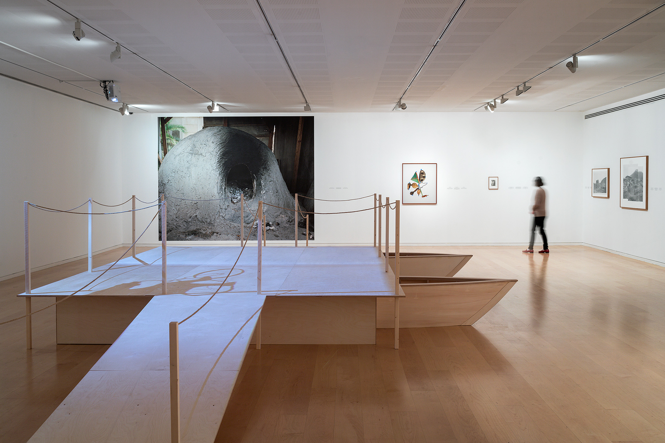 Installation view of museum gallery space with a large wood sculpture- a ramp elevated by two small boats - in the center and works (photographs) in the background - of the Frena, a bread baking oven, and a photographic collage of an amulet.