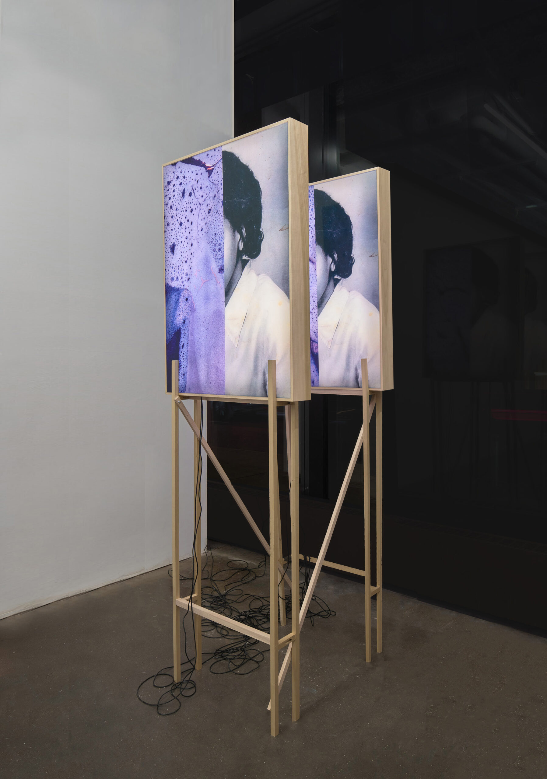 A structure containing two light boxes, each featuring the same image of a woman whose face is partially obscured by a print of a ceramic scan. Wires descend from the light boxes and coil on the floor beneath.