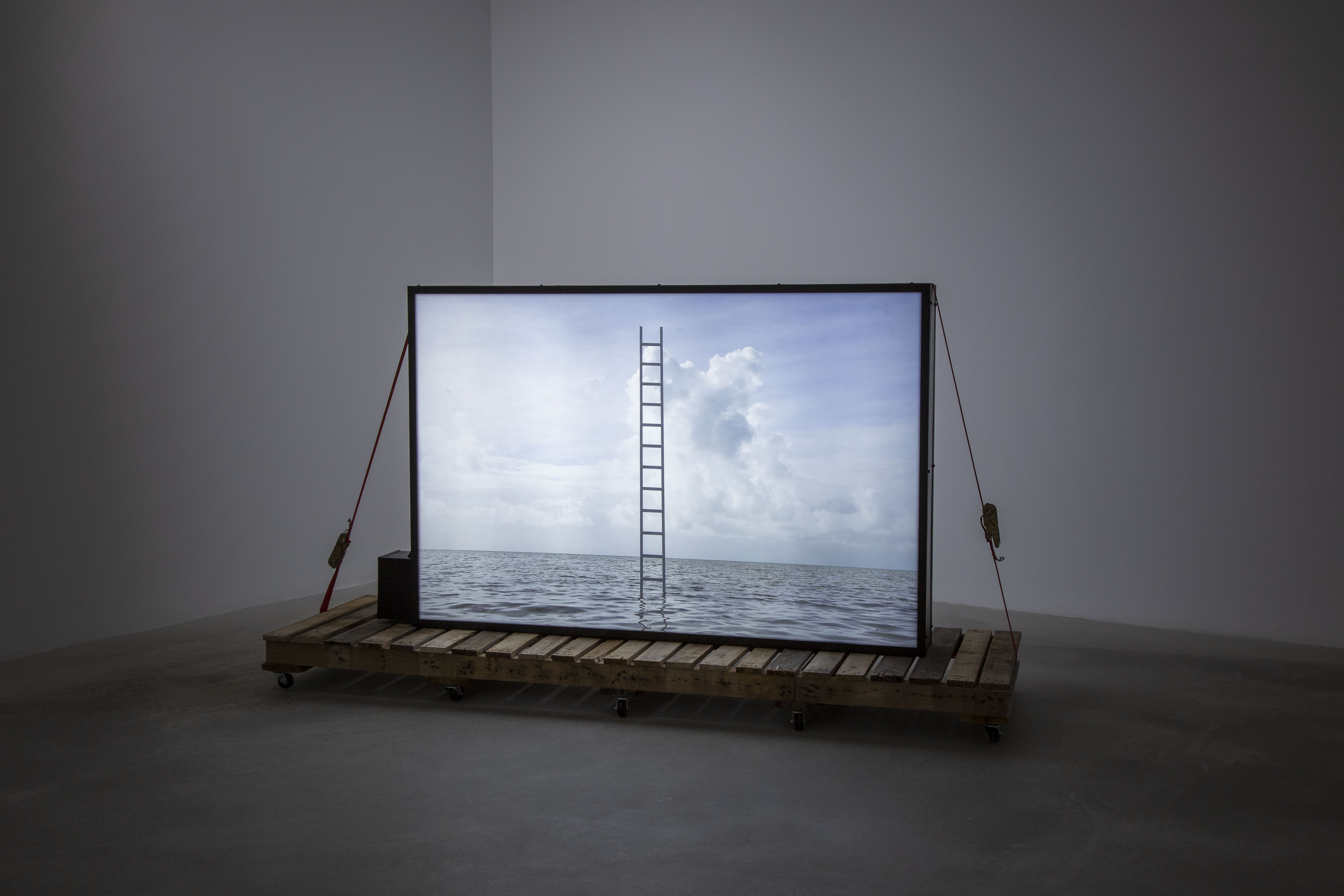 An industrial light box, 5 ft wide, features an image of a ladder protruding from the sea. The scene is idyllic with the bright blue sky, fluffy white clouds. The lightbox is held down on a rudimentary cart like structure on wheels with a ratchet strap.