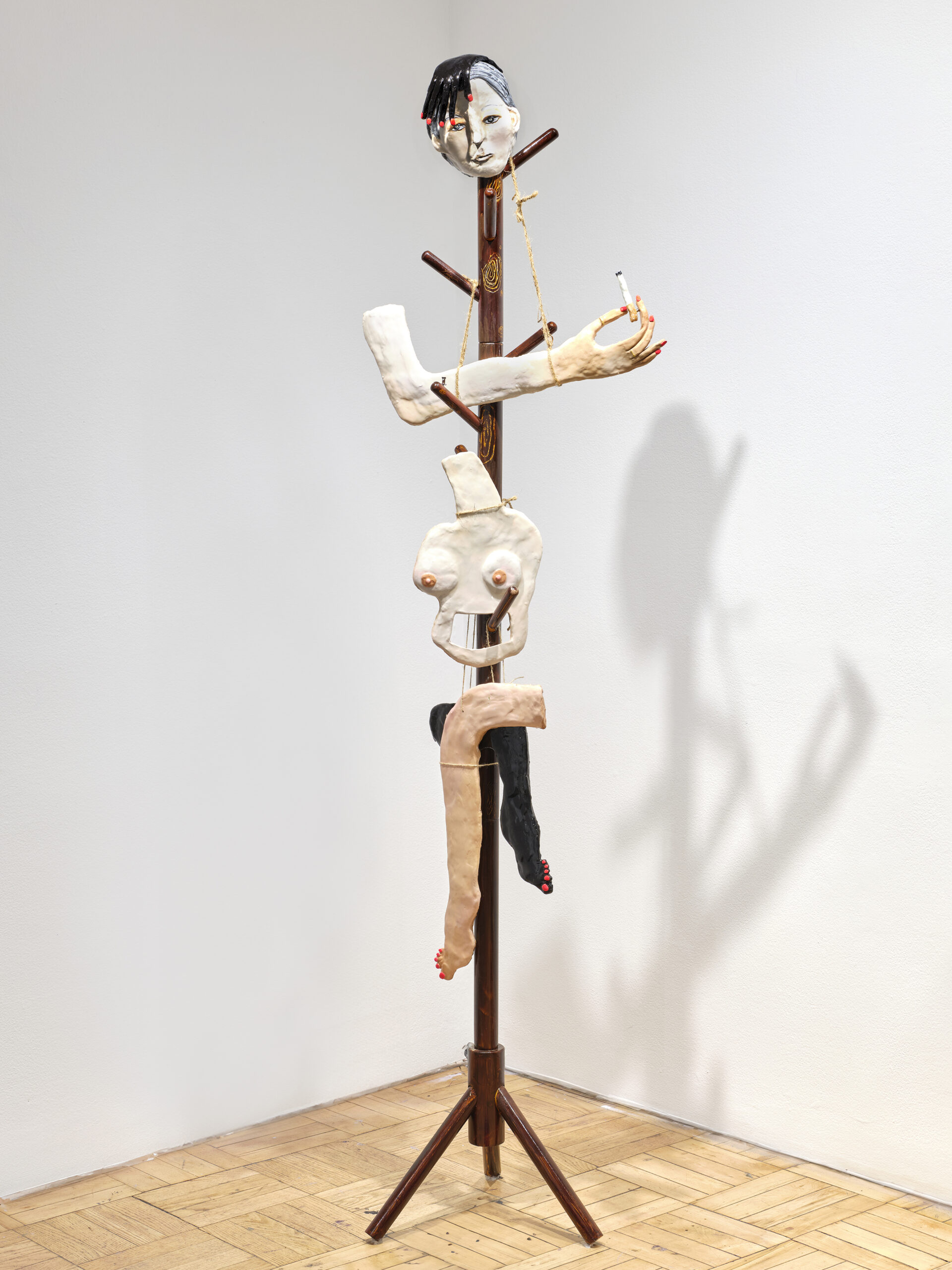 Figurative sculpture constructed from a wooden coat rack adorned with hand-sculpted elements, including a detailed face, arms, torso, and legs. One arm extends outward, holding a cigarette, while the legs, one black and one pale, dangle from the lower section