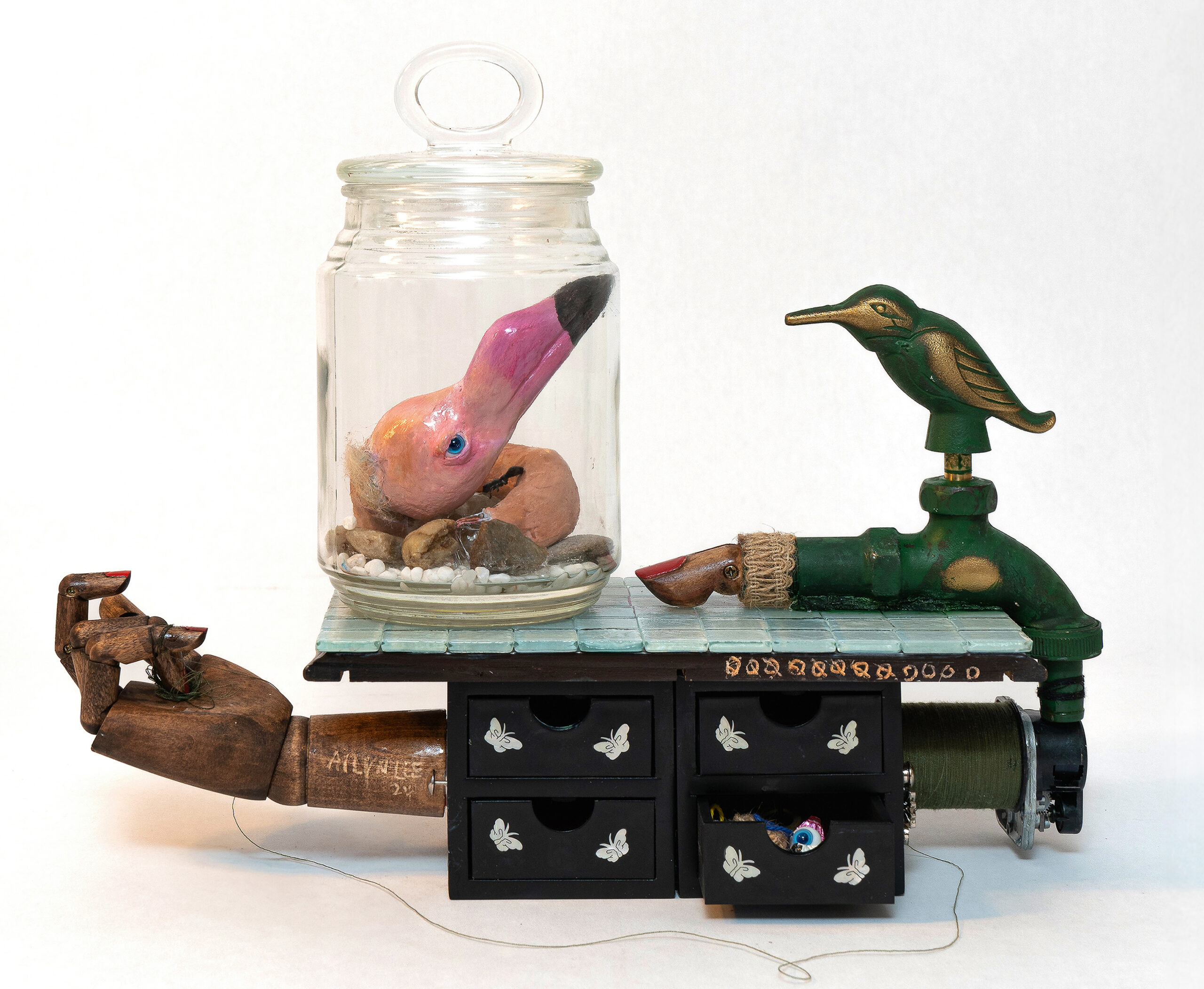 Surreal assemblage sculpture featuring a glass jar containing a pink bird-like head resting on stones, placed atop a small black chest of drawers adorned with butterfly motifs. A wooden hand extends from the side, holding a delicate thread, while a green faucet with a golden bird figure emerges on the opposite end.