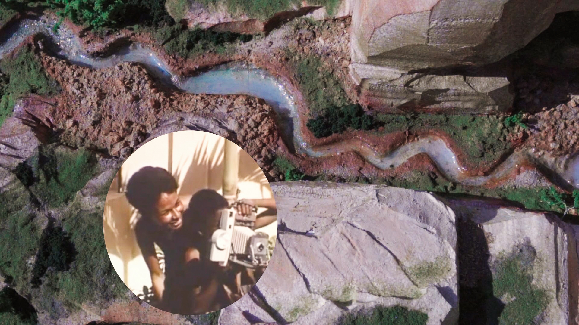 A textured diorama of a winding river cutting through rocky terrain, with mossy green patches suggesting vegetation. A circular inset in the lower left corner features a vintage scene of two Black filmmakers operating a camera, evoking themes of archival memory and historical storytelling.