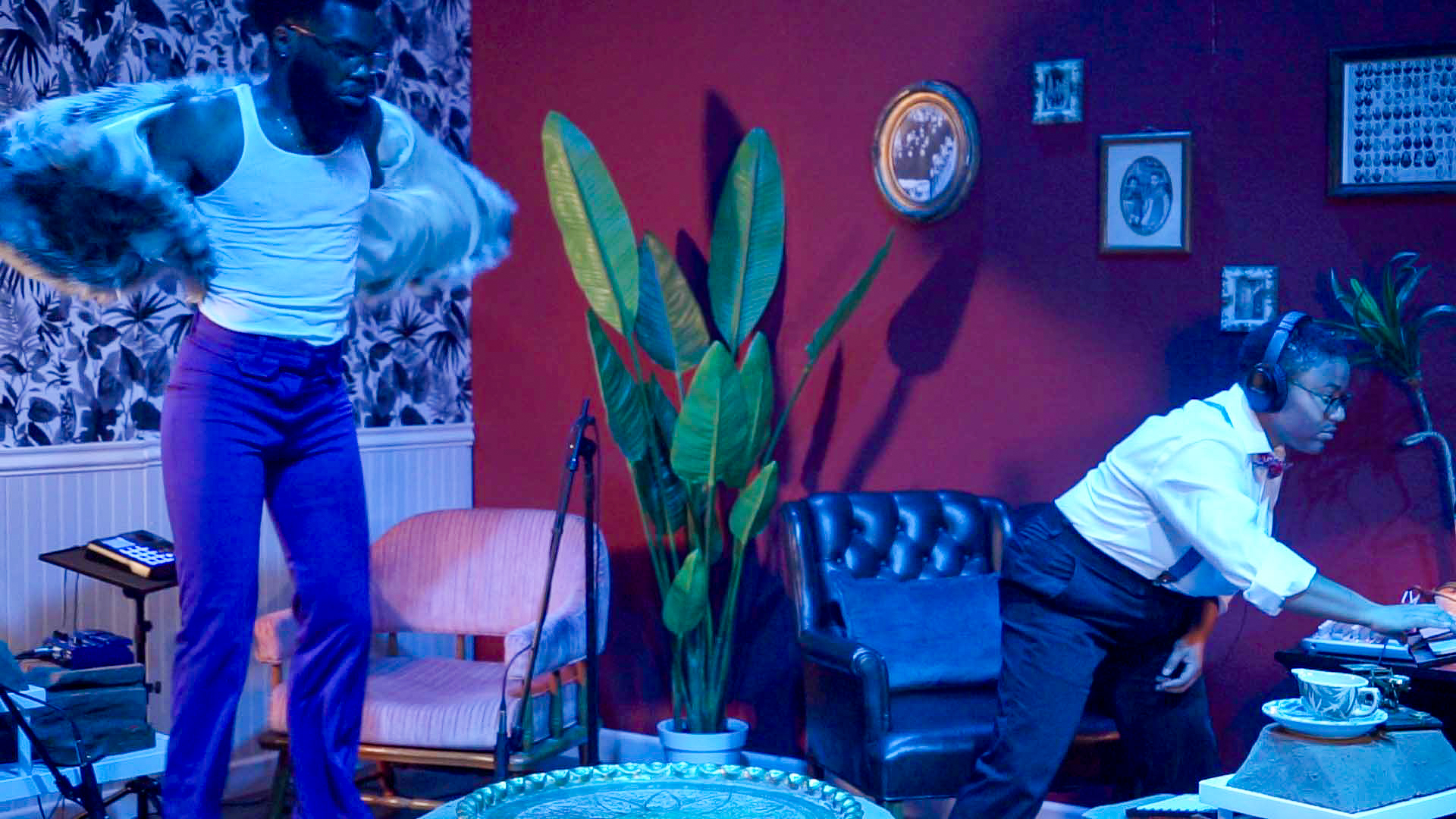 A dimly lit, vibrant interior bathed in deep blue and red hues. A person in a white tank top and purple pants energetically removes a faux fur jacket, while another, dressed in suspenders and a bow tie, focuses intently on a DJ setup. The retro-styled room is adorned with vintage portraits, lush plants, and eclectic furniture, suggesting a theatrical performance or cinematic reenactment.