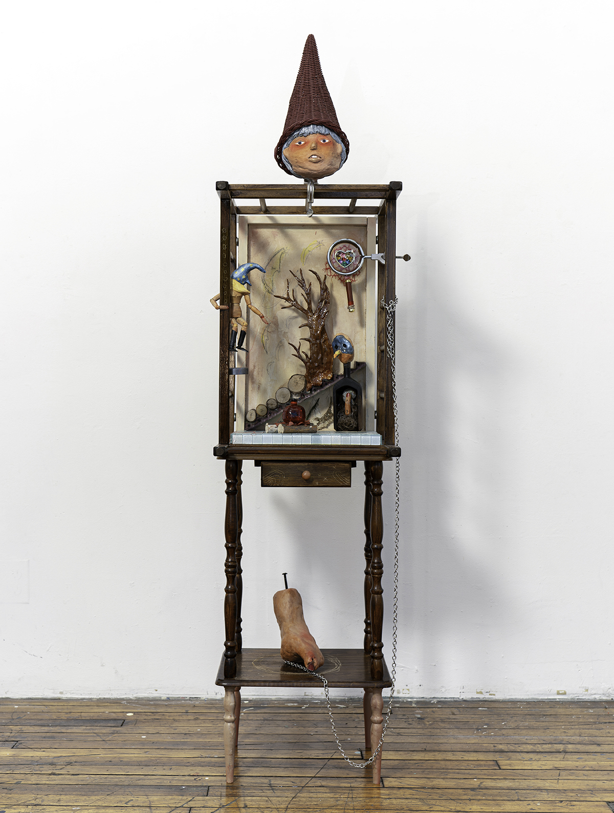 Assemblage sculpture on a wooden stand, featuring a doll-like head in a red hat, miniature figures, a barren tree, and chained objects.