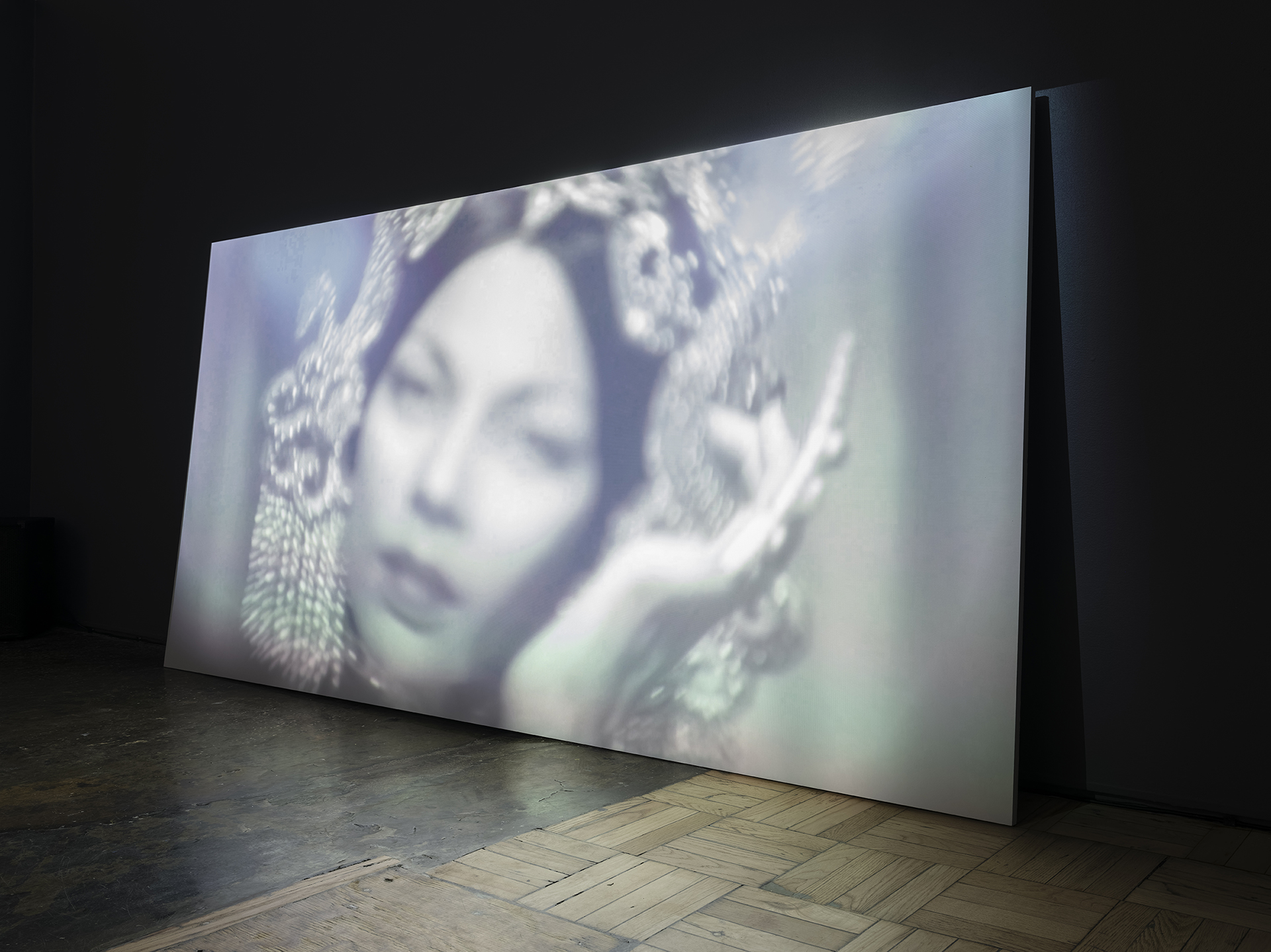 A large screen placed against the wall showing a still from Silhouettes Remain of a woman's face in black and white.