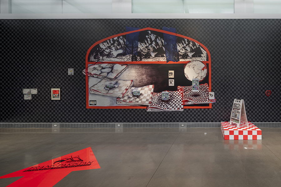 A large red-outlined vinyl collage spans a checkered wall, featuring drawn maps, black mountains, and a globe. Nearby, a checkered pedestal holds a signboard.