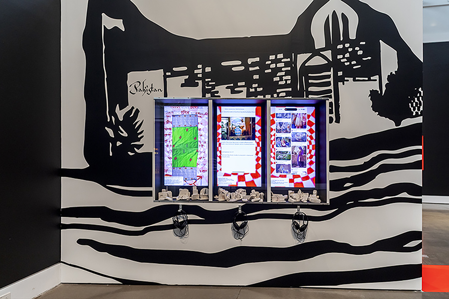 Interactive exhibit featuring three digital screens displaying colorful images and maps. Black and white mural background with headphones below.