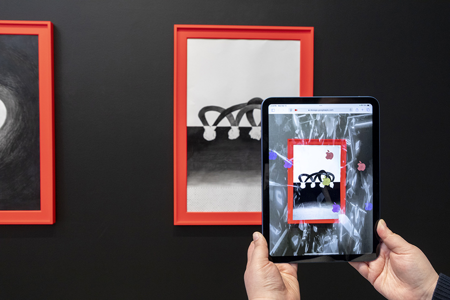 Hands hold a tablet displaying digital art of a red-framed black-and-white drawing, mirroring an adjacent digital augmented image. Modern, interactive exhibition theme.