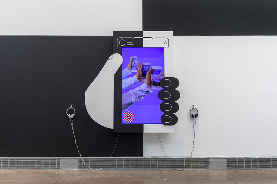 An art piece shows a large hand holding a smartphone with a screen displaying multiple hands with phones. Headphones hang on a black and white checkered wall.