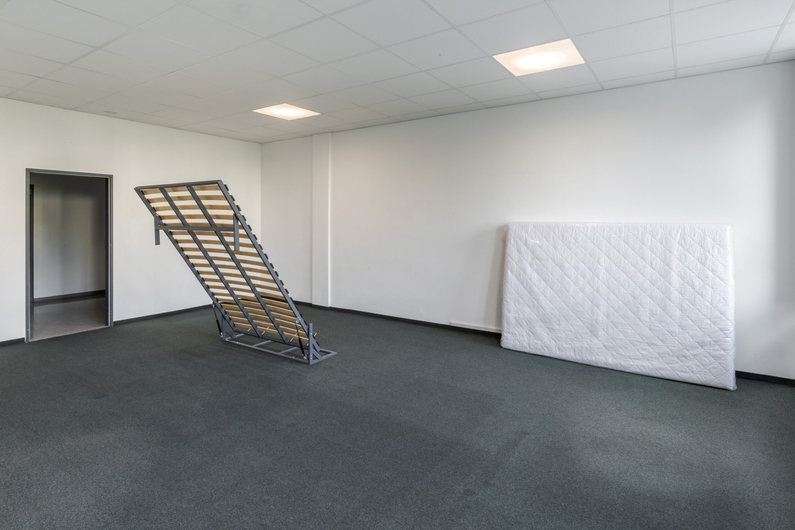 The frame of a grey metal Murphy wall bed stands at an angle, half open half closed in the middle of an office room. Adjacent in the room leaning against the wall is a white double mattress wrapped in plastic.