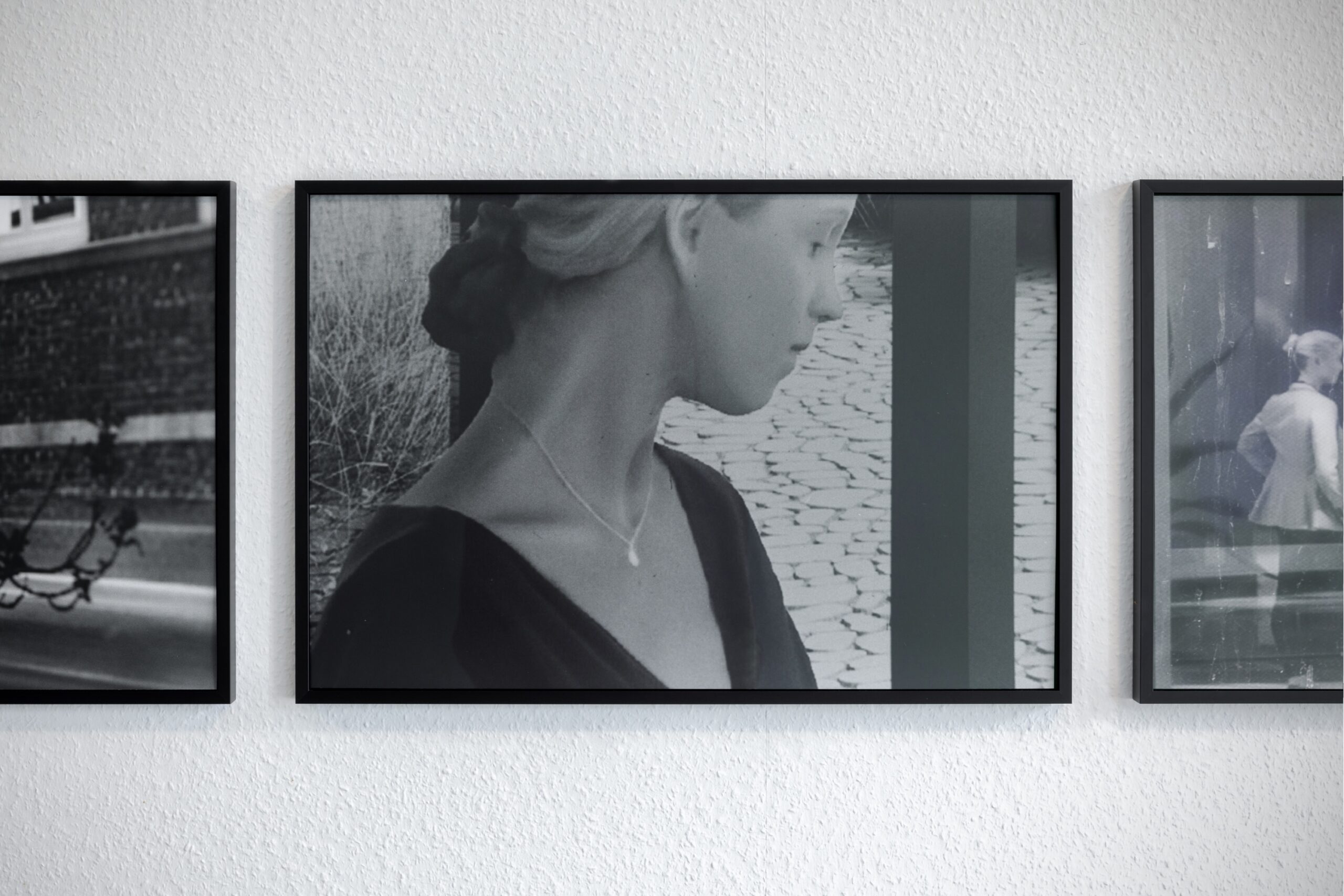A framed black and white silver gelatine print of a woman's profile. She is looking away from the camera, her features feel uncanny, as if computer generated. The framed image is flanked by framed photographs in a similar style with just the edge of each image visible.