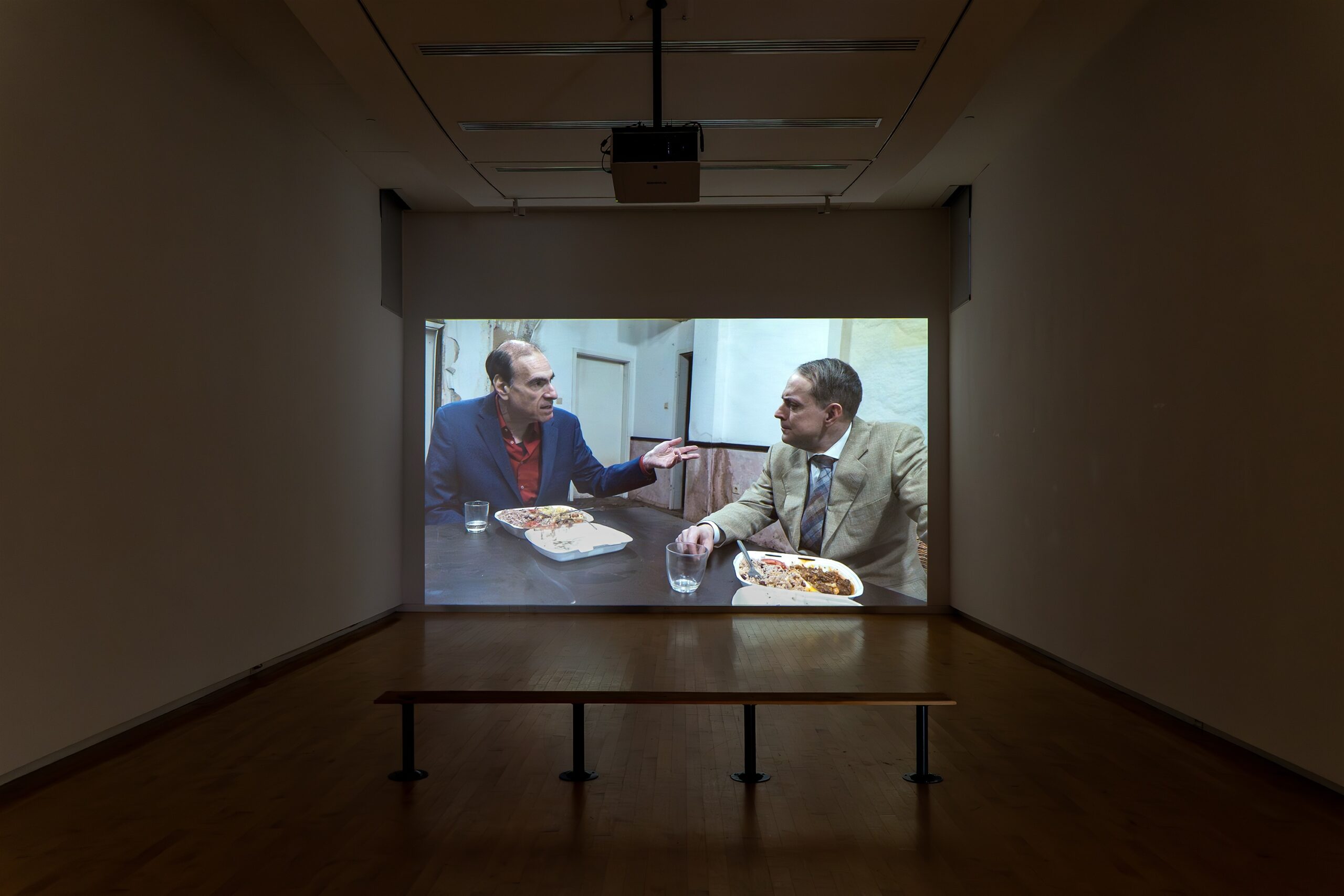 Two men in navy and beige suits sit at a table in the dilapidated shell of a building. The wallpaper is peeling off the walls and there are damp stains rising from the ground. The men eat food from takeaway cartons with drink glasses of champagne.